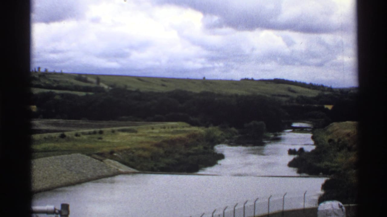 1972 North Dakota 8mm frame showing a river, grassy banks, distant hills and cloudy sky from an elevated viewpoint.