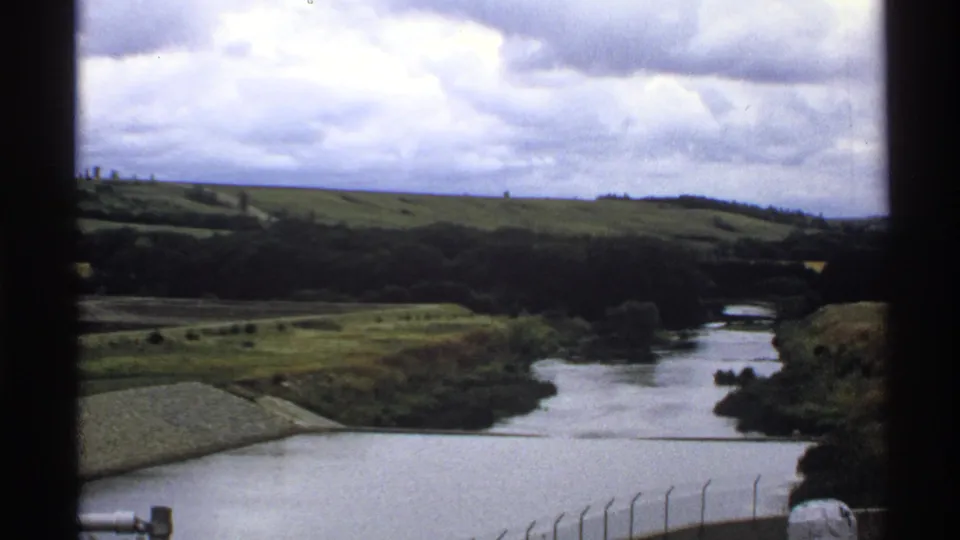 1972 North Dakota 8mm frame showing a river, grassy banks, distant hills and cloudy sky from an elevated viewpoint.