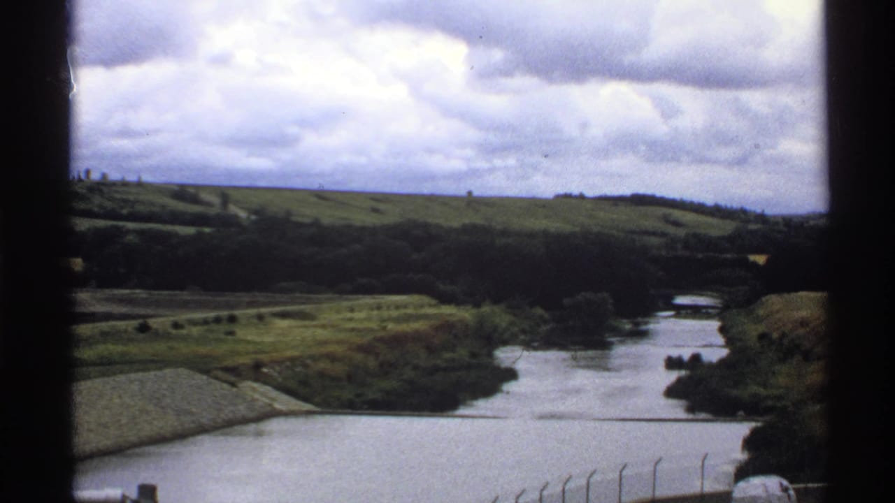 North Dakota river landscape on 8mm film, with rolling grass, distant trees, cloudy sky and a small water-control structure visible.
