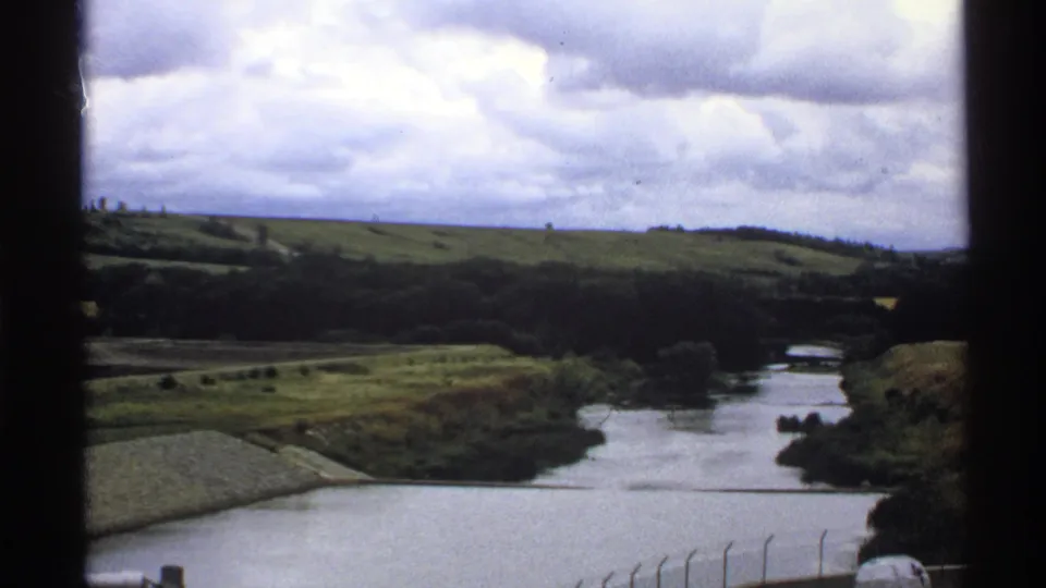 North Dakota river landscape on 8mm film, with rolling grass, distant trees, cloudy sky and a small water-control structure visible.