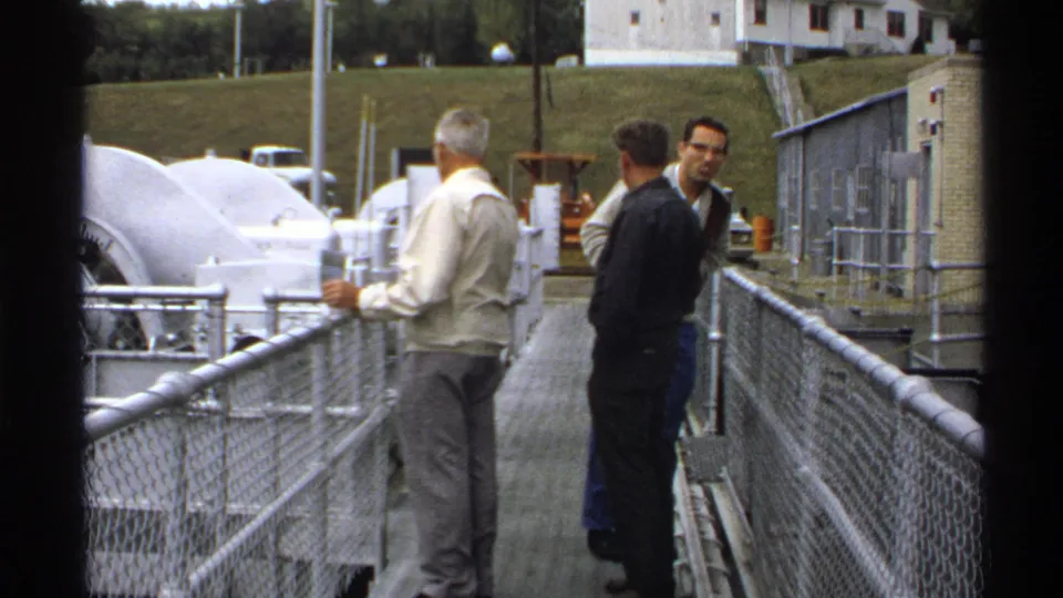 1972 North Dakota 8mm frame of three senior men on a fenced metal walkway beside large covered industrial equipment.