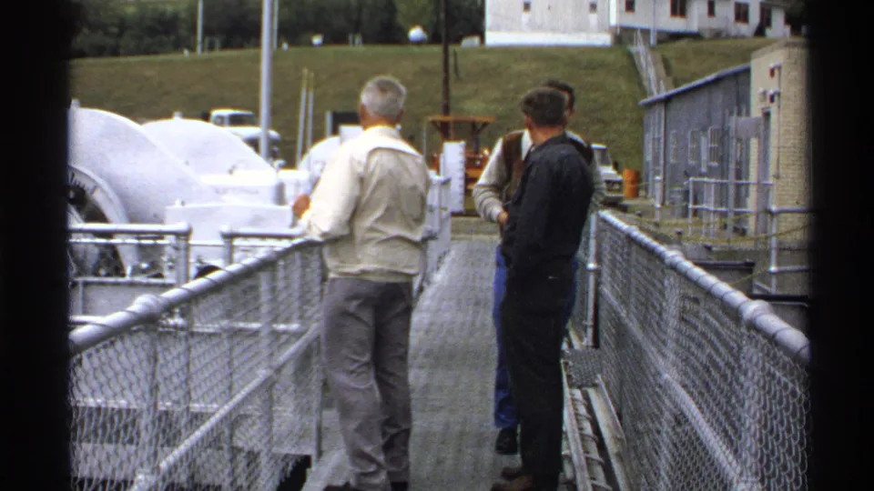 Senior men in casual 1970s clothing observe large facility equipment from a chain-link walkway in North Dakota.