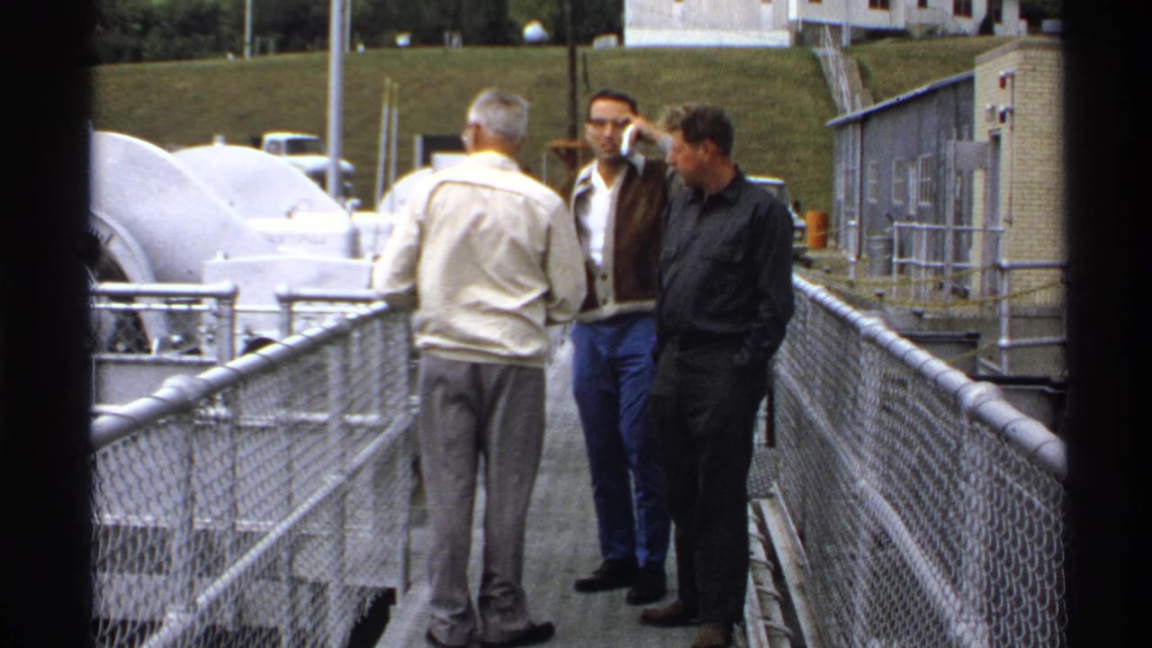 Three adult men converse on a metal walkway at a North Dakota industrial facility in 1972, with white machinery and a grassy hill behind them.