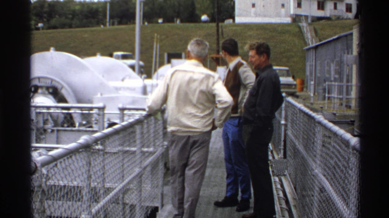 Three men face one another on a metal walkway near railings and industrial machinery.