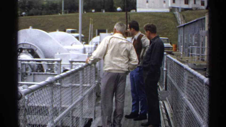 Three adult men remain on the walkway at the industrial site, seen near chain-link fencing, white machinery, and a low utility building.