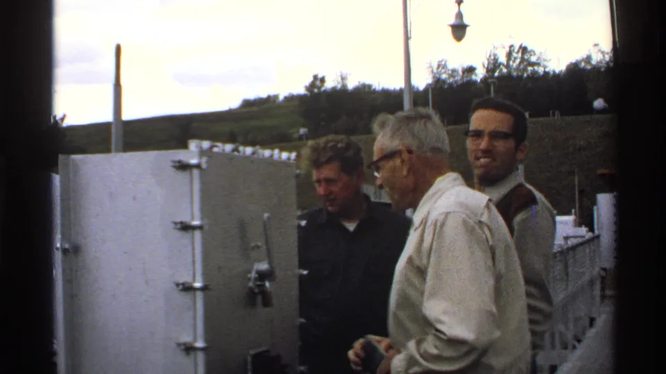 A closer 1972 North Dakota view shows the three men near a large metal enclosure, with the industrial site and hillside behind them.