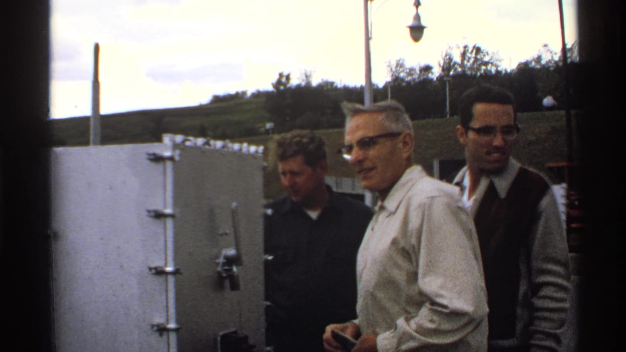 Three adult men stand near large buildings and utility poles in a rural setting.