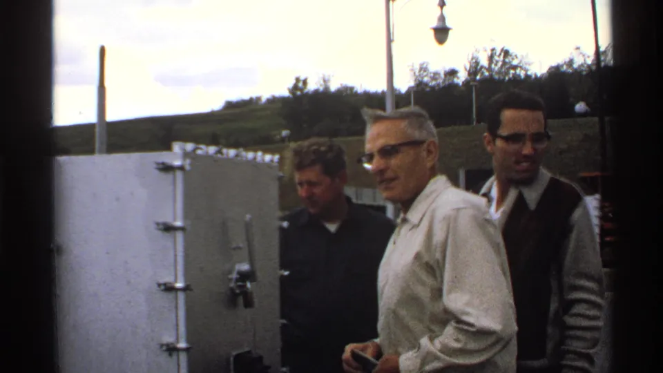 In North Dakota 1972 home-movie footage, three adult men stand near a facility with utility poles, grassy ground, and low buildings.