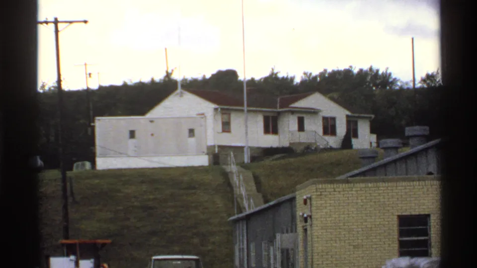A white rural building sits on a grassy North Dakota hillside, with utility poles and low facility structures in view.