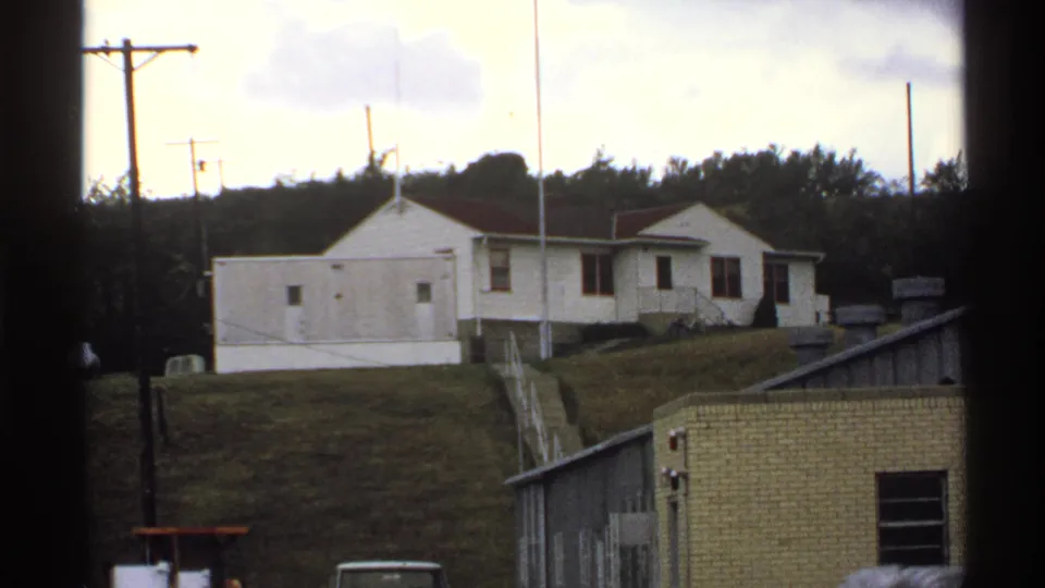 The frame shows rural North Dakota facility buildings, a white structure on a hill, utility poles, and a low brick building in the foreground.