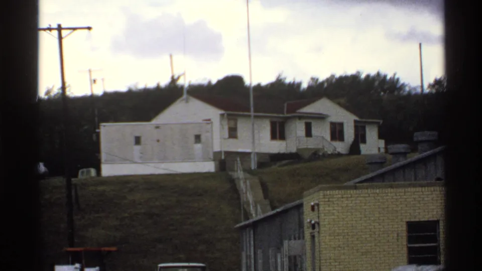 A white building and utility poles stand on a grassy North Dakota hillside, with a low brick structure and trees visible nearby.