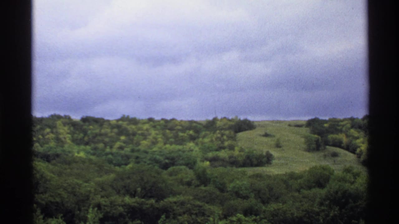1972 North Dakota 8mm frame showing wooded rolling hills under a cloudy sky with dark scan borders.