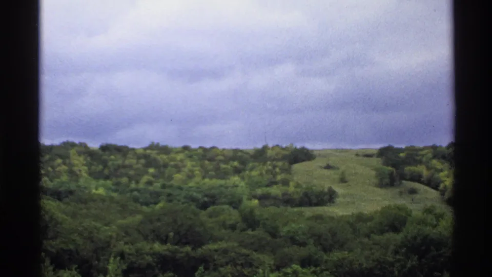 1972 North Dakota 8mm frame showing wooded rolling hills under a cloudy sky with dark scan borders.
