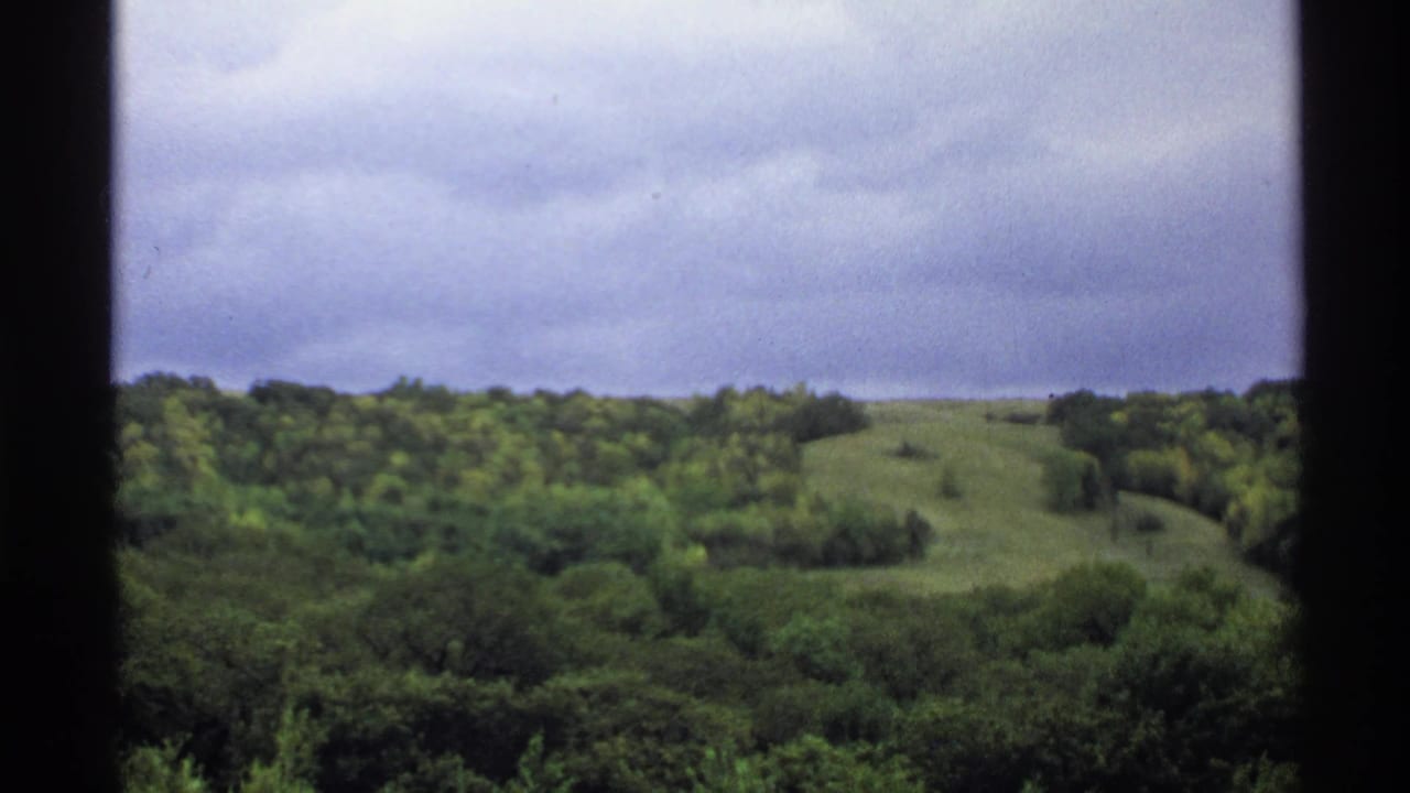 1972 North Dakota landscape frame with a grassy hill rising behind dense trees beneath an overcast sky.