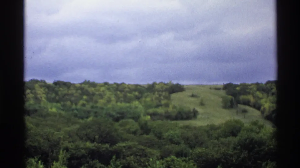 1972 North Dakota landscape frame with a grassy hill rising behind dense trees beneath an overcast sky.