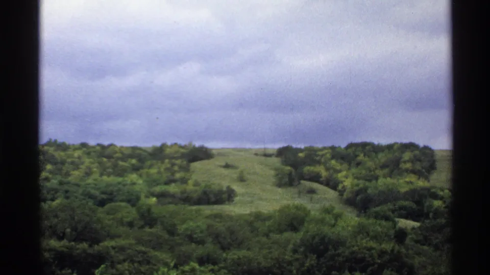 1972 North Dakota home-movie frame with a green hill, surrounding woodland, clouded sky, and visible film gate edges.