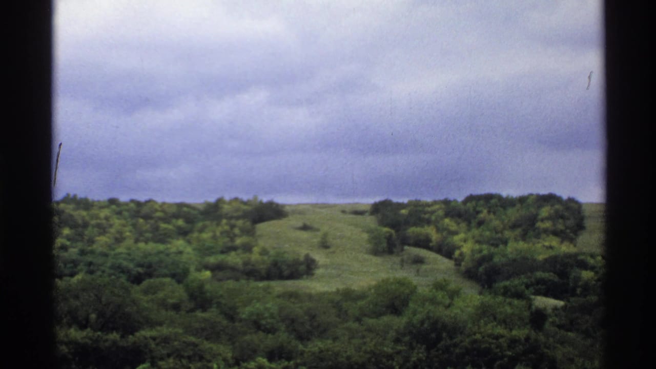 1972 North Dakota home-movie view of green hills and trees, framed by dark window edges beneath an overcast sky.