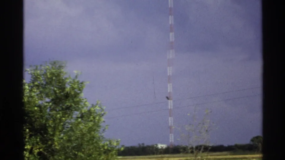 Wide home-movie view of the KTHI-TV Tower in rural North Dakota, with trees in the foreground and a cloudy blue-gray sky behind it.