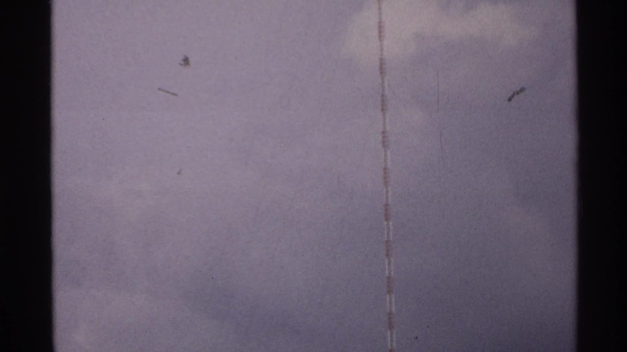 1972 North Dakota home-movie frame with a striped tower edge and faint dark specks against an overcast sky.
