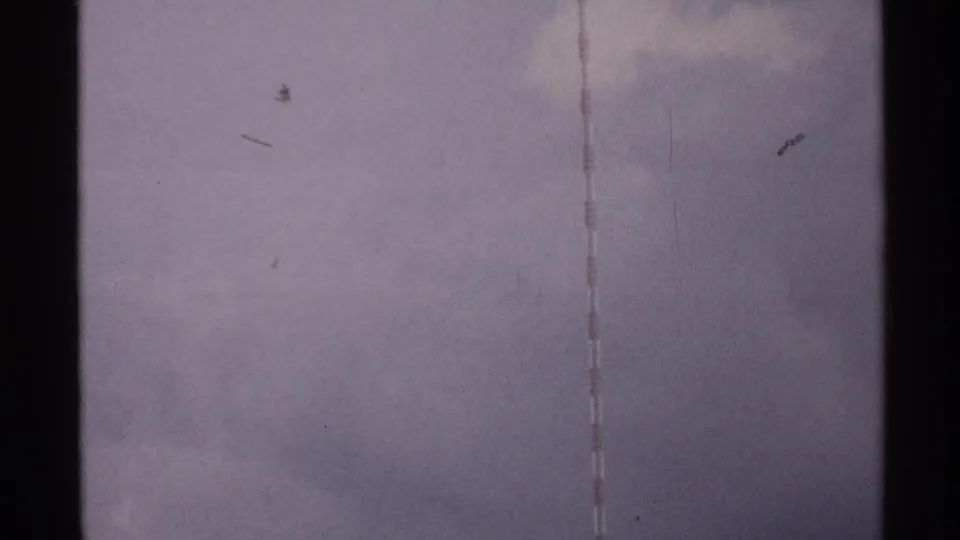 1972 North Dakota home-movie frame with a striped tower edge and faint dark specks against an overcast sky.