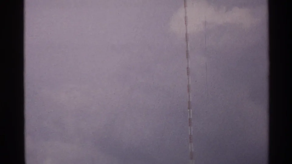 Faded 8mm frame from North Dakota in 1972 showing a slim striped tower line against hazy clouds.