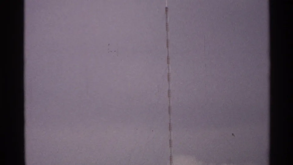 1972 North Dakota 8mm frame showing dark vertical tower-like forms and faint lines against a pale, grainy sky.