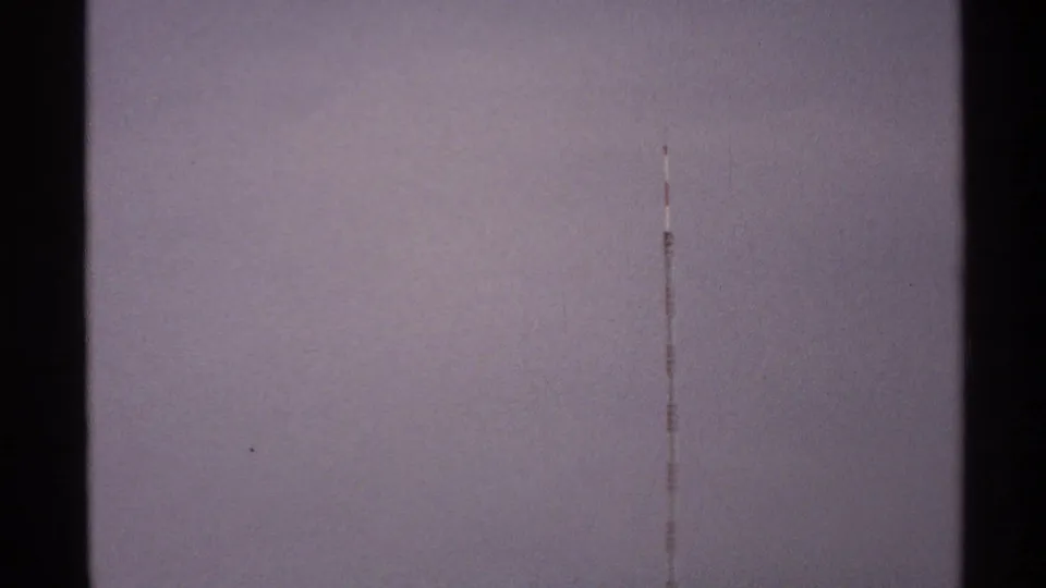 A dark vertical tower-like form and a thin line stand against a pale grainy sky in this 1972 North Dakota 8mm frame.