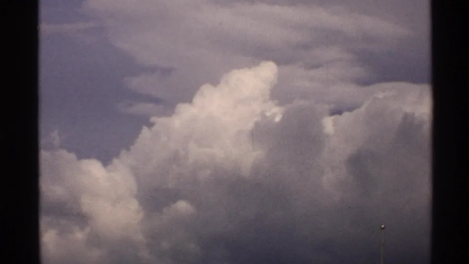 1972 North Dakota 8mm frame showing billowing cumulus clouds with shadowed bases under a subdued blue sky.