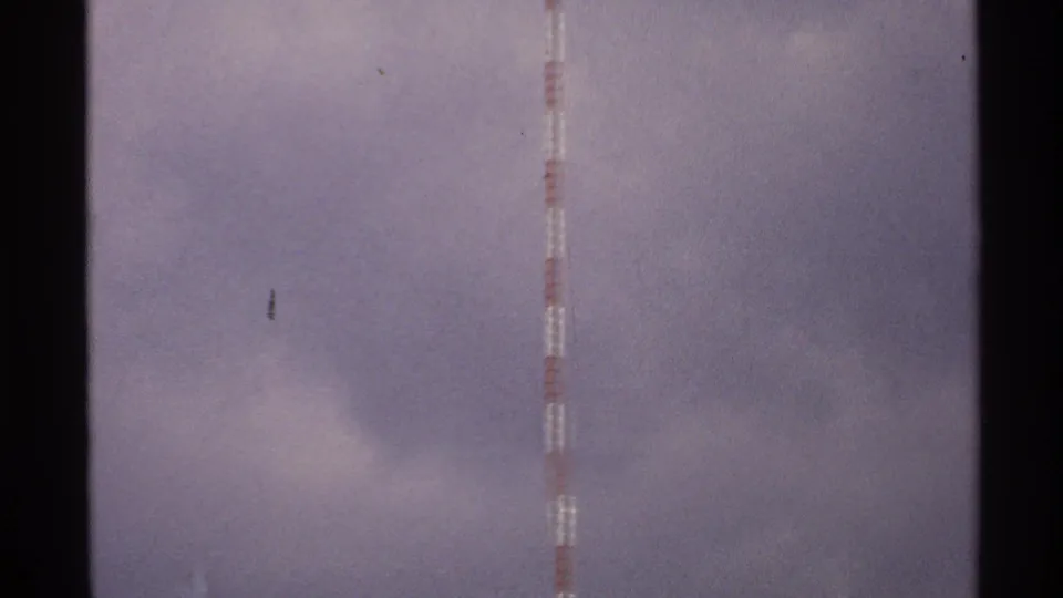 A lower section of the striped tower appears near the frame edge, with cloudy sky and visible 8mm film texture.