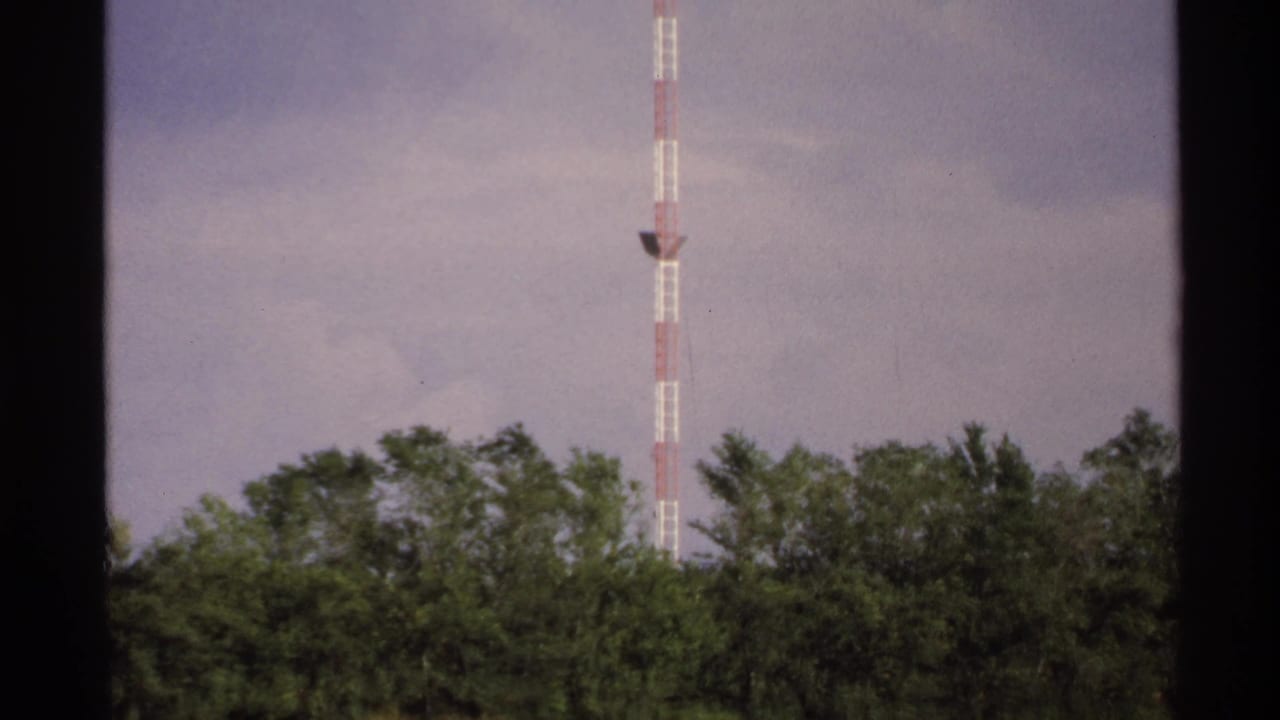 1972 8mm frame showing a tall red-and-white communication tower above trees, filmed from a moving roadside viewpoint.