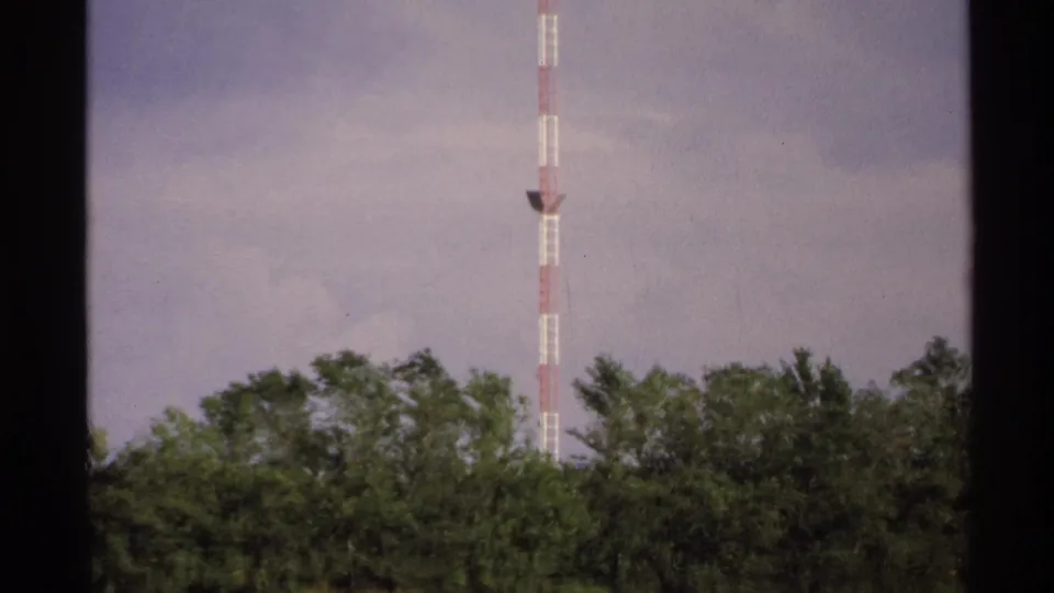 1972 8mm frame showing a tall red-and-white communication tower above trees, filmed from a moving roadside viewpoint.