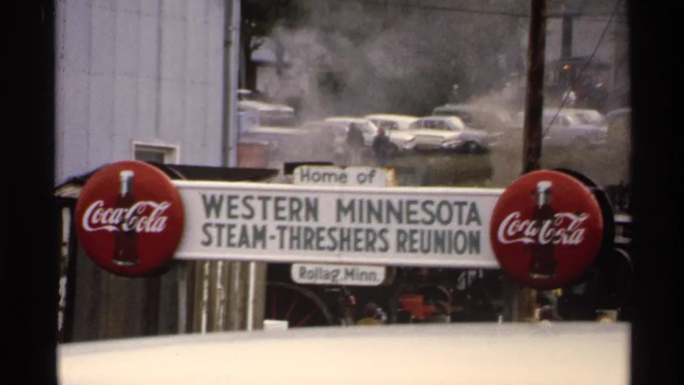 1970s 8mm frame of the Western Minnesota Steam-Threshers Reunion sign, flanked by Coca-Cola logos, with steam and classic cars in back.