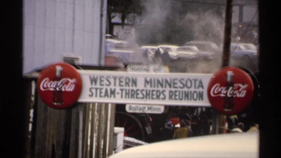 Closing 1972 frame of the Rollag reunion sign with Coca-Cola logos, drifting steam, parked cars, and distant visitors near the entrance.