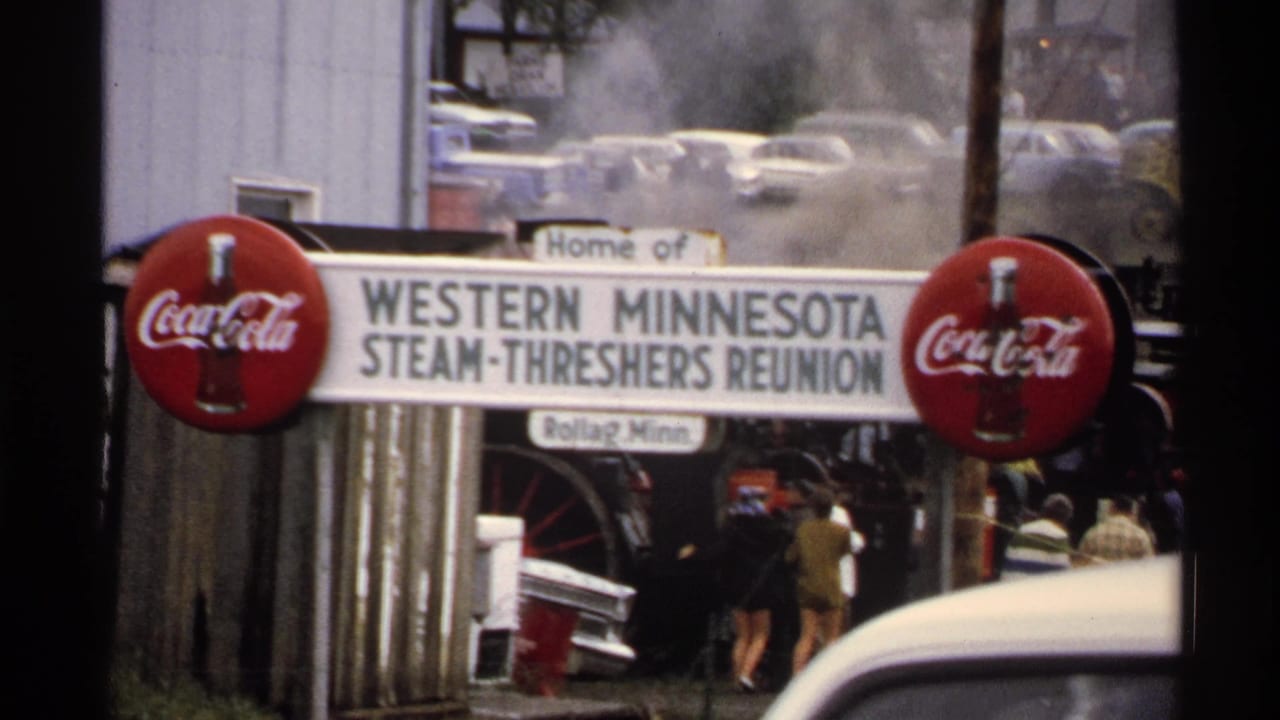1972 8mm frame showing the Western Minnesota Steam-Threshers Reunion sign at Rollag, with Coca-Cola signs, steam, cars, and visitors in the background.