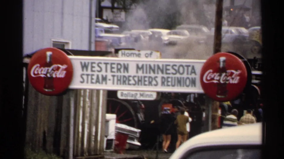 1972 8mm frame showing the Western Minnesota Steam-Threshers Reunion sign at Rollag, with Coca-Cola signs, steam, cars, and visitors in the background.