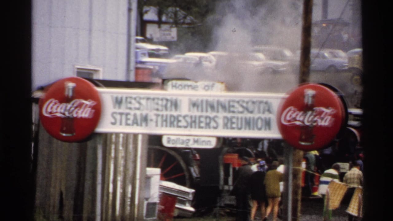 1972 8mm frame of the reunion entrance sign and Coca-Cola branding, with steam, trees, parked cars, and fairgoers near equipment.