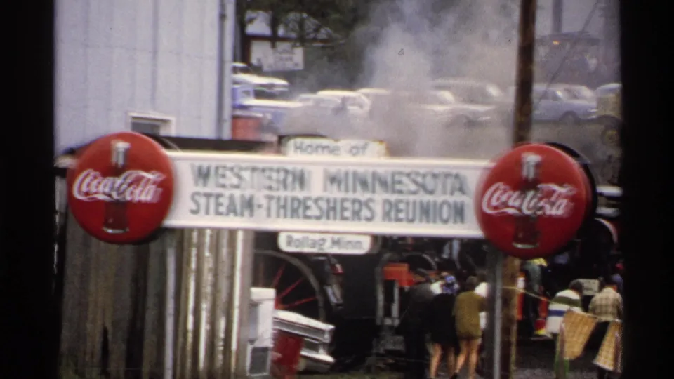 1972 8mm frame of the reunion entrance sign and Coca-Cola branding, with steam, trees, parked cars, and fairgoers near equipment.