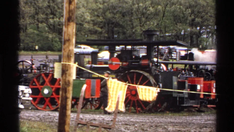 1972 8mm frame showing a black steam traction engine with red wheels at the reunion grounds, partly viewed through poles and a rope barrier.