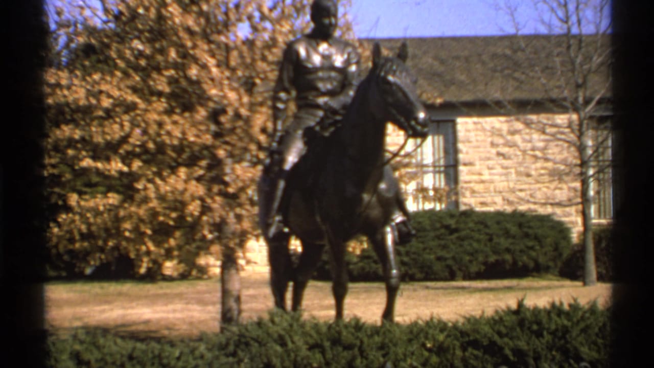 Bronze equestrian statue seen through dark foreground framing near a stone building and autumn trees.