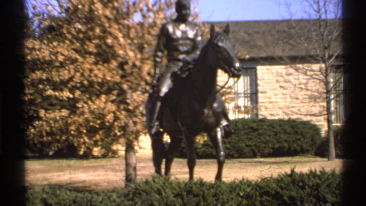 Equestrian statue in a park setting with shrubs, yellow leaves, and a stone building behind it.