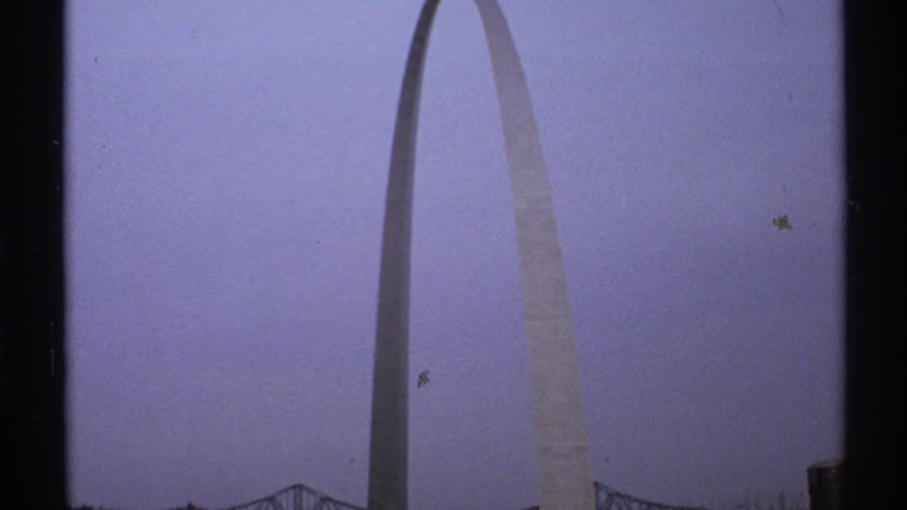 1972 Saint Louis home-movie frame showing the Gateway Arch from a distance against a cloudy pale sky, with a bridge visible near the lower edge.
