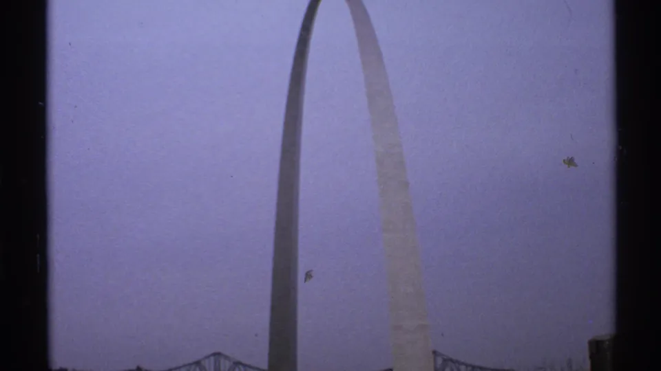 1972 Saint Louis home-movie frame showing the Gateway Arch from a distance against a cloudy pale sky, with a bridge visible near the lower edge.