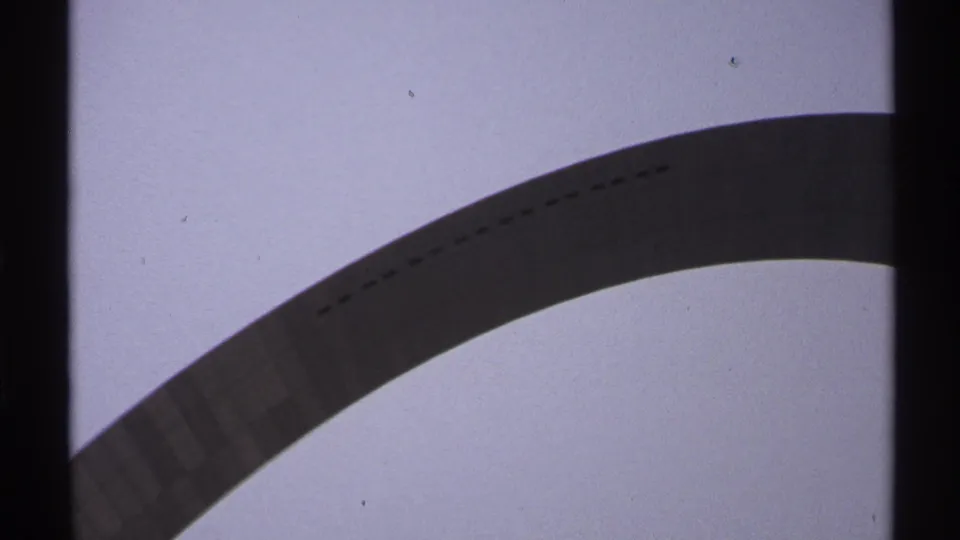 1972 Saint Louis home-movie frame cropped close on the Gateway Arch curve, showing its dark underside against a pale cloudy sky.