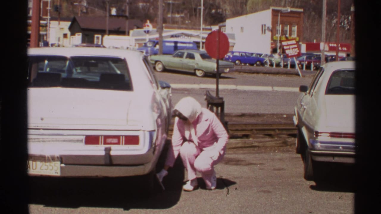 Woman in a pink outfit crouching beside a white car in a parking lot.