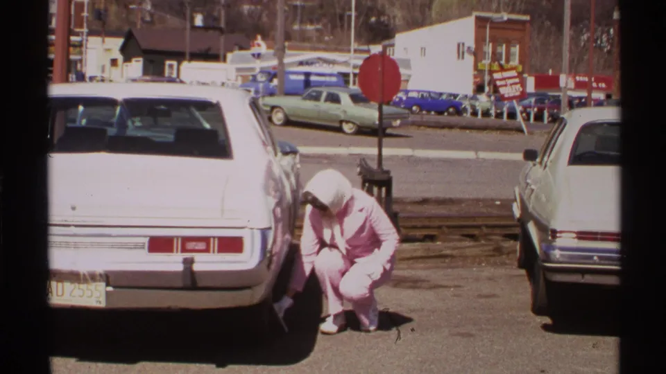 In 1972 Saint Louis footage, a woman in a pink pant suit crouches by a white car while checking a tire near parked cars and storefronts.