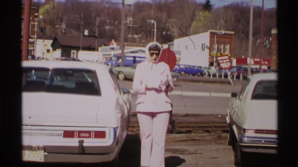 A woman in a pink pant suit and scarf stands between parked cars in 1972 Saint Louis, with a stop sign and storefronts behind her.