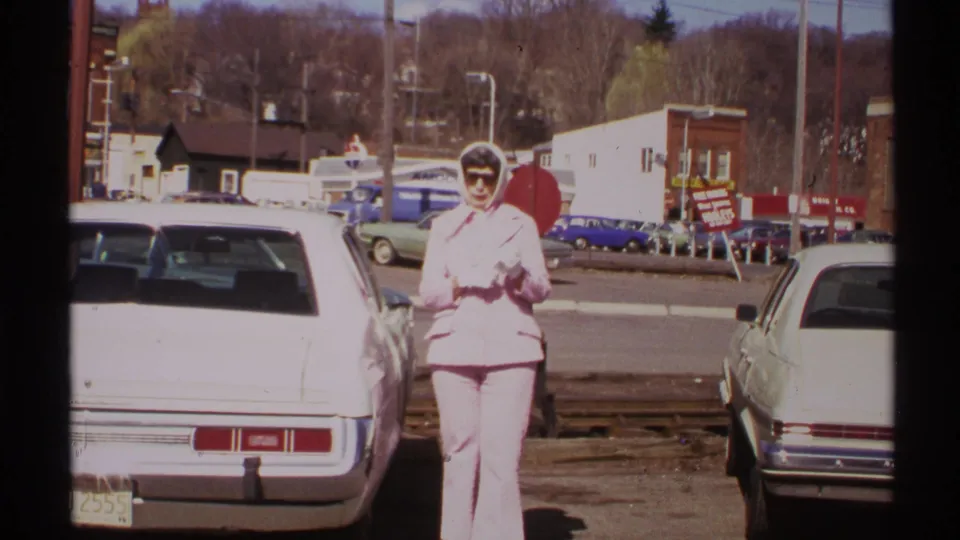 The woman stands in the parking lot near the white car, framed by parked vehicles, a stop sign, and storefronts in 1972 Saint Louis.