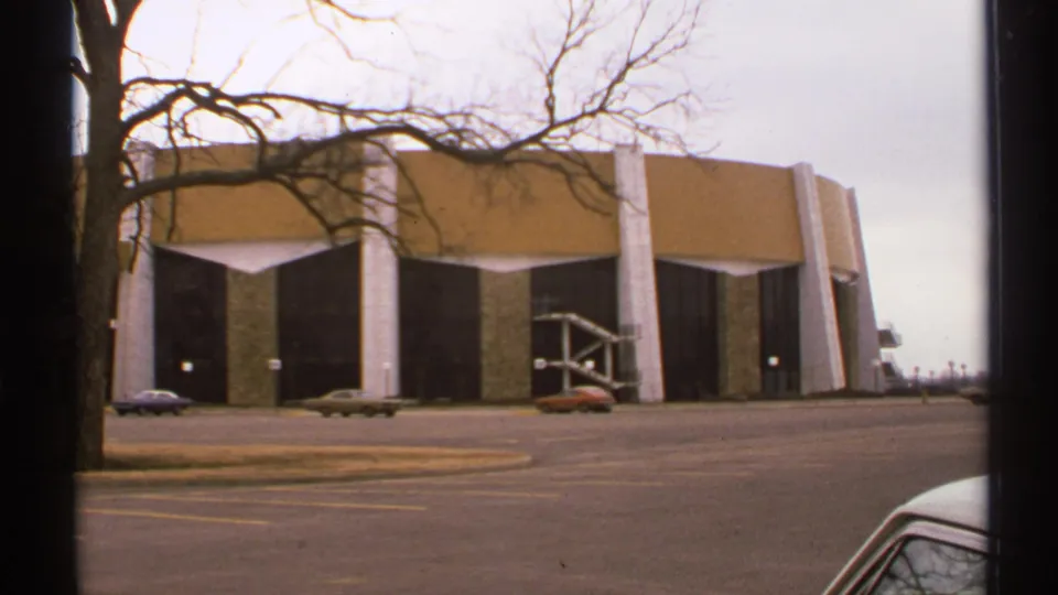 1972 Tulsa 8mm frame showing a large modernist building with white supports, tan upper panels, dark bays, bare trees, and parked cars.