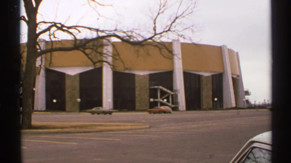 1972 Tulsa home-movie frame of a broad modernist structure with angled white columns, tan panels, dark recessed areas, and cars in the foreground.