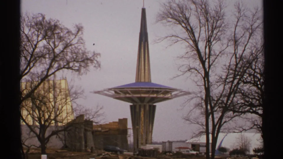 1972 Tulsa 8mm frame of a tall pointed tower rising from a disc-shaped platform, framed by leafless trees and low structures below.